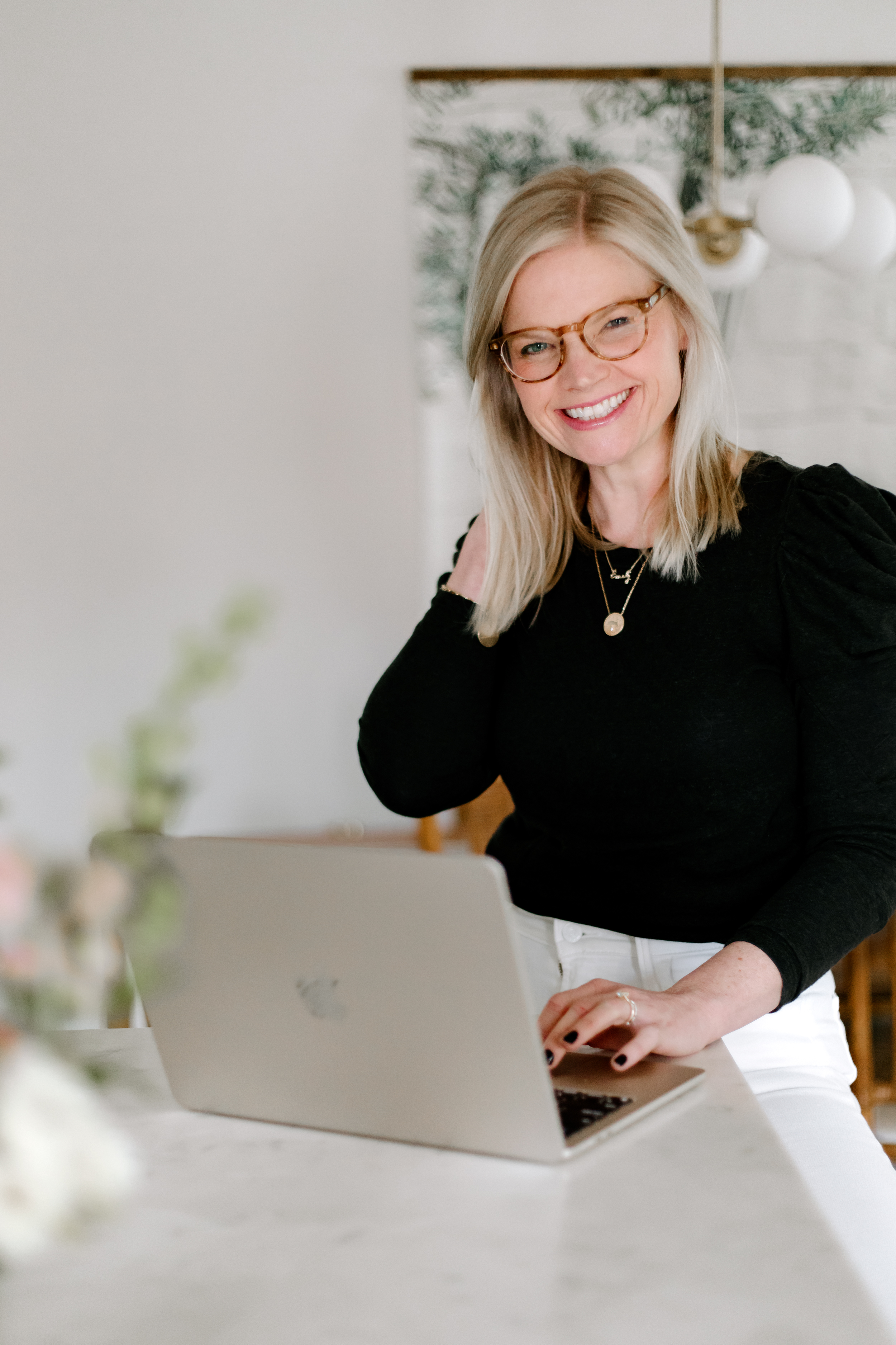 Em wearing glasses, a black sweater and white pants, smiling behind her laptop