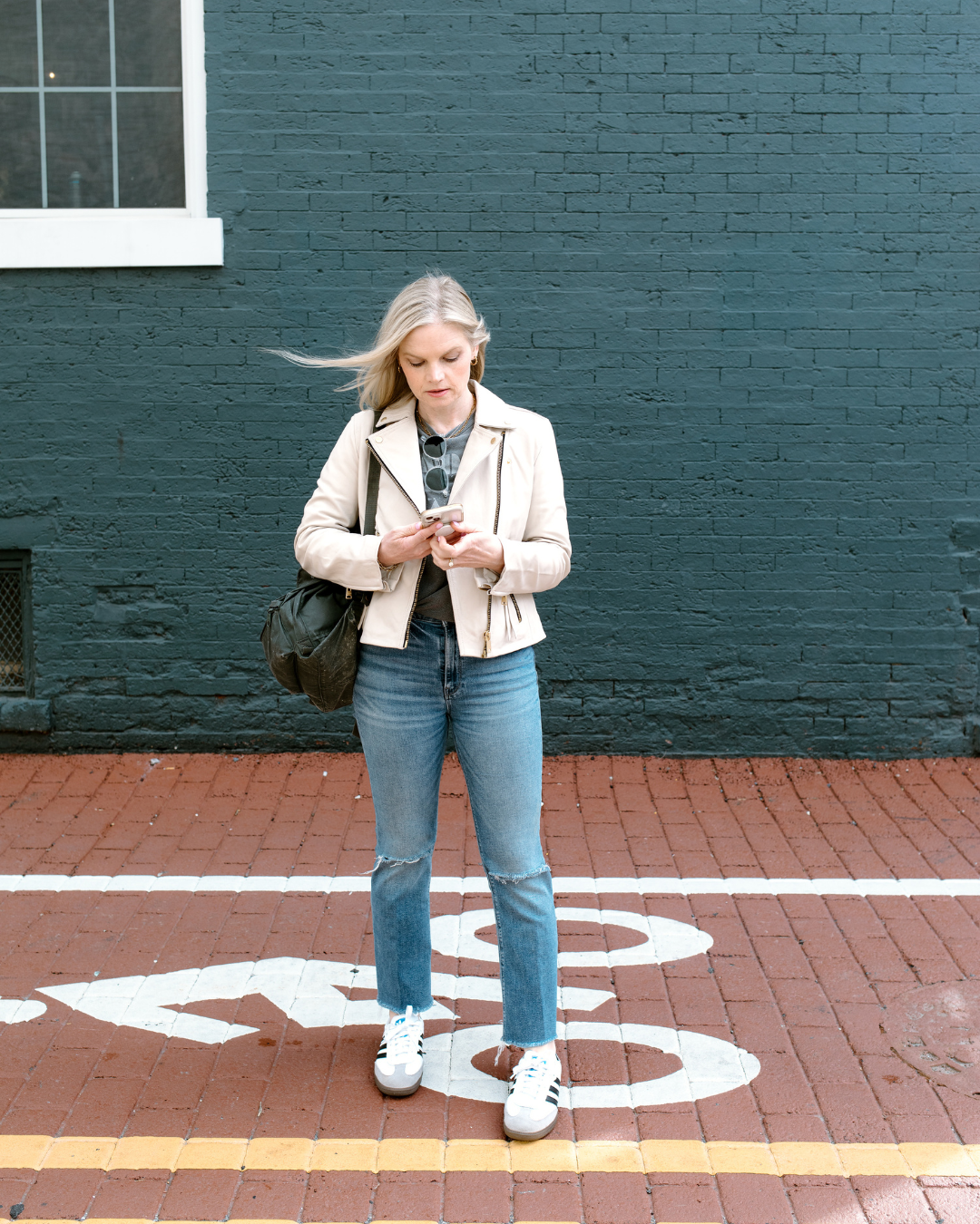 Em standing outside with a white leather jacket and jeans on, looking down at her phone