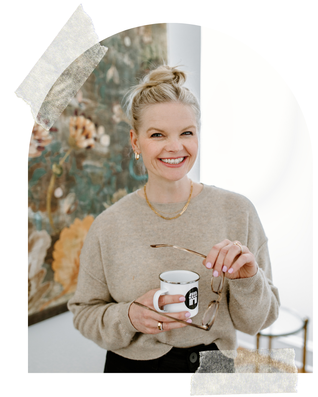 Em holding her glasses and coffee mug and smiling at the camera wearing a tan sweater