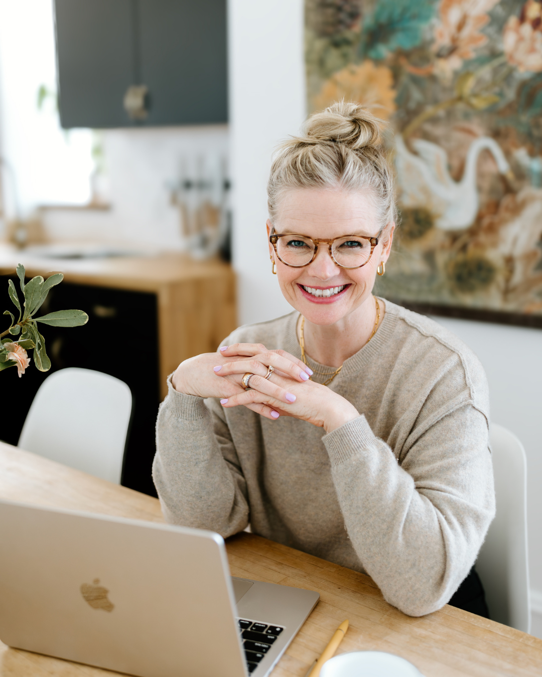 Em wearing a tan sweater and her glasses, smiling behind her laptop