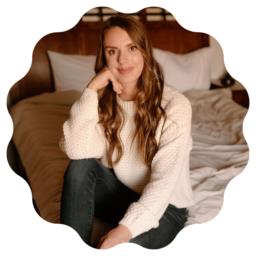 woman in a cream sweater sitting on a bed with her hand under her chin, smiling at the camera