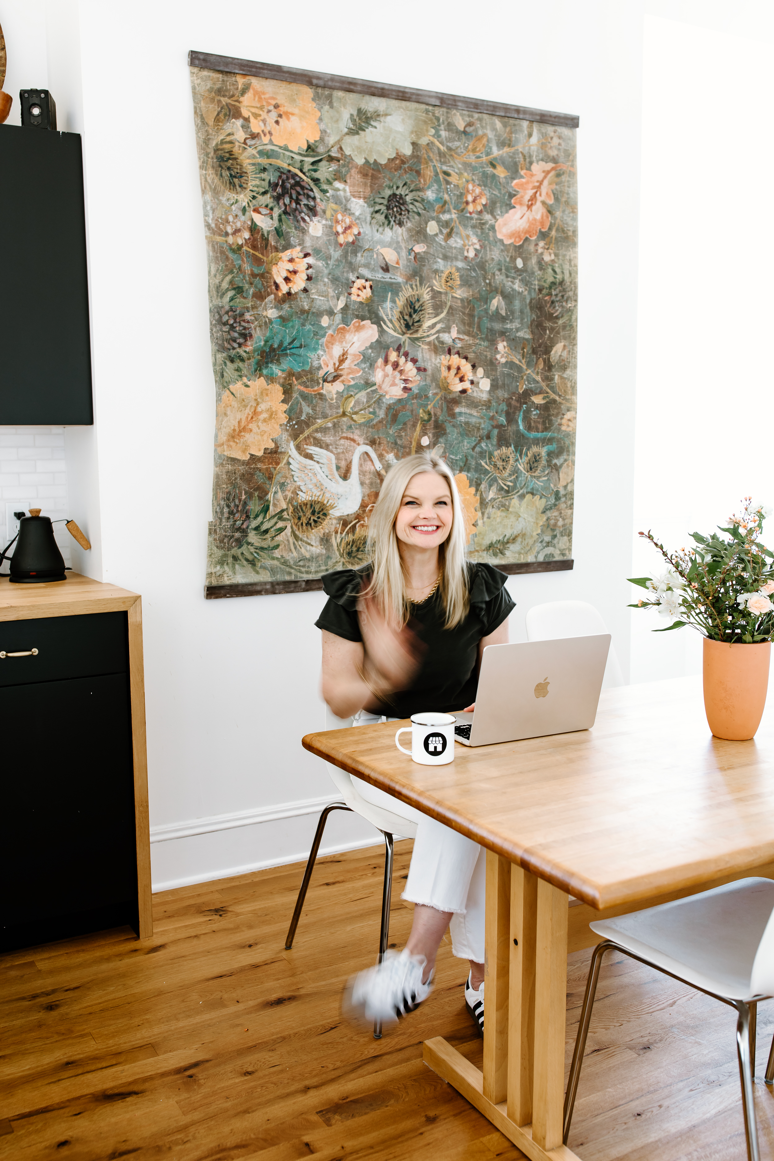 Em wearing a grey shirt sitting behind her laptop waving, with her coffee mug beside her and a floral print on the wall behind her
