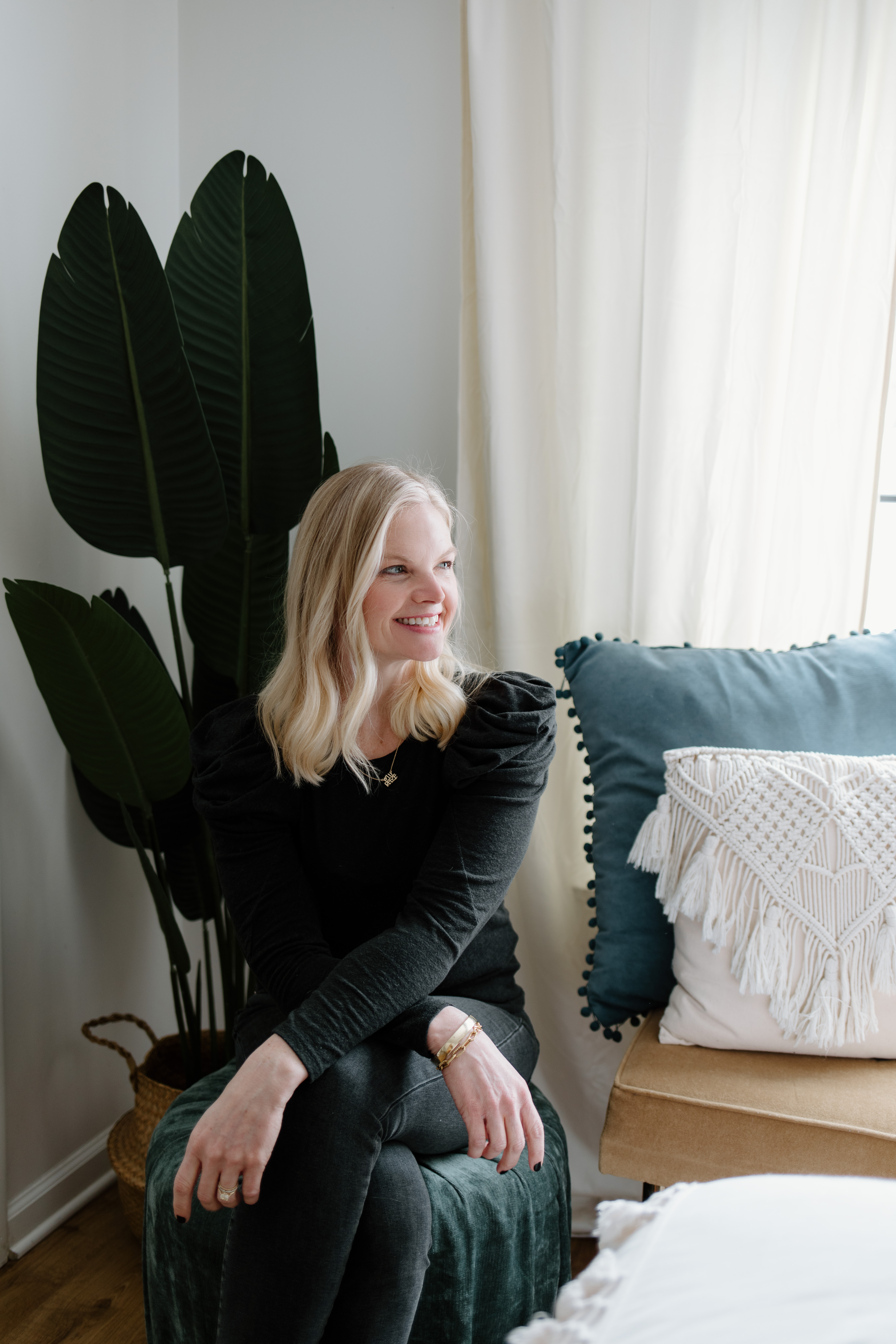 Em wearing a black long sleeve shirt, sitting on a chair next to a blue and white pillow, smiling off to the side